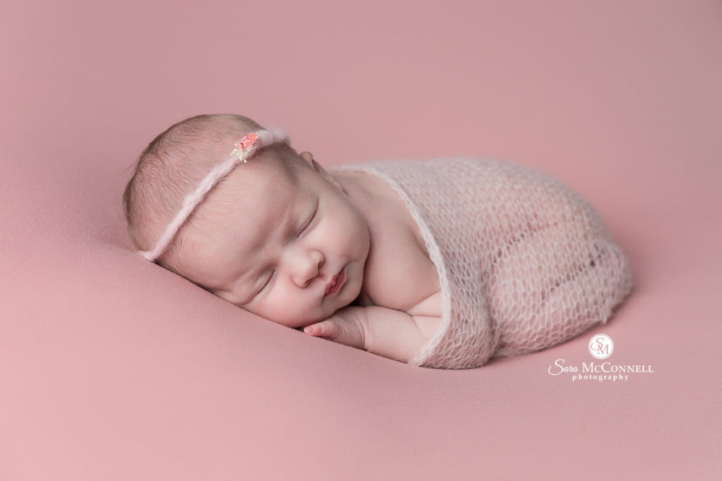 baby girl wrapped in a knit fabric sleeping on a pink backdrop