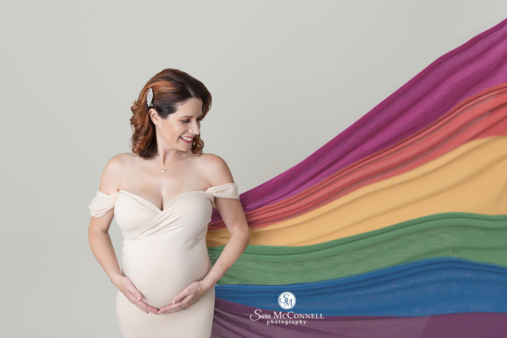 expectant mother cupping her belly as a rainbow of material is behind her