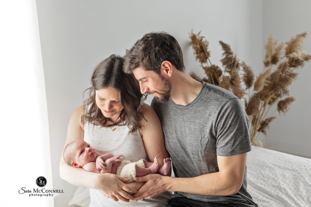 Photo of a mom and dad holding their newborn while they look down at them. Newborn photographers ottawa.
