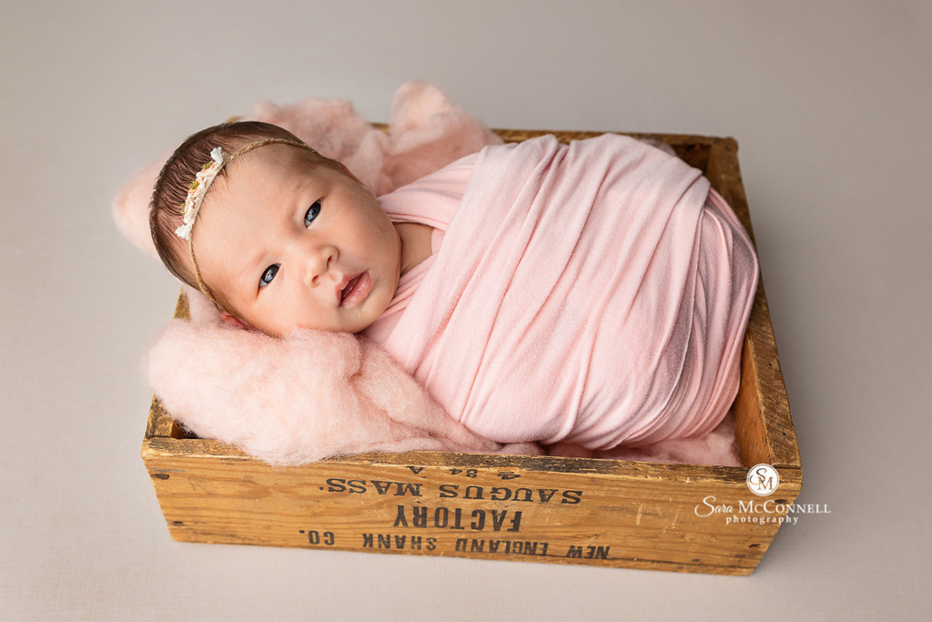 Photo of a newborn baby girl posed in a wooden prop in a blog post called "What if my baby is awake?" Maternity photography near me.